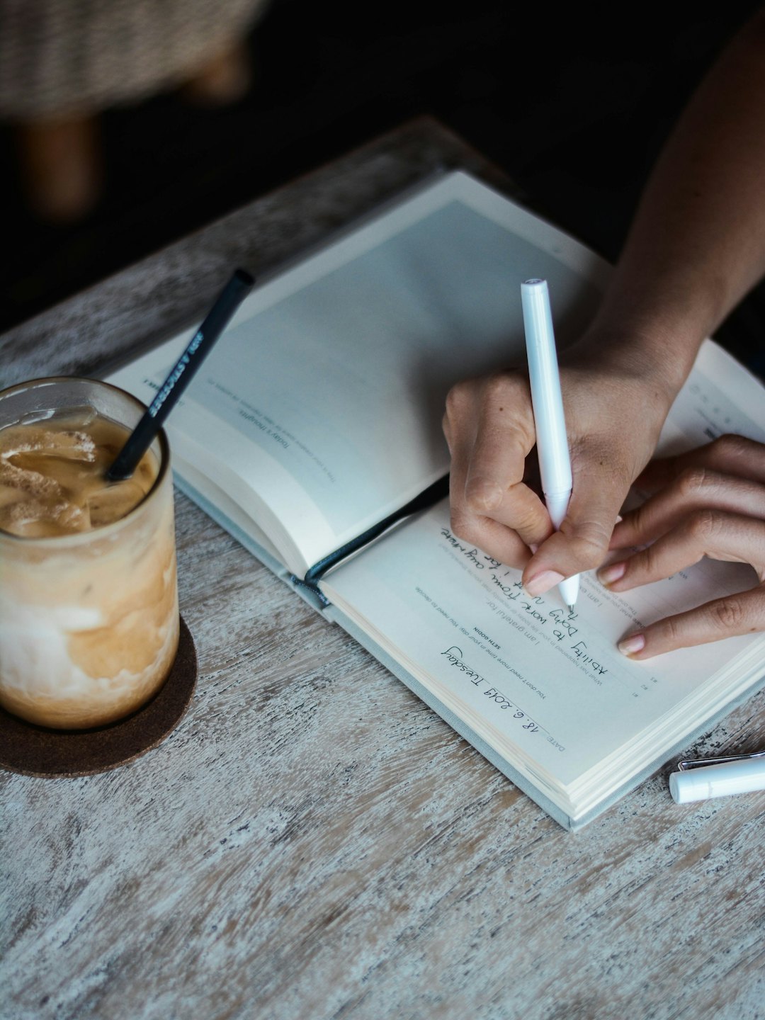 Hands writing in a journal with a white marker and iced coffee.